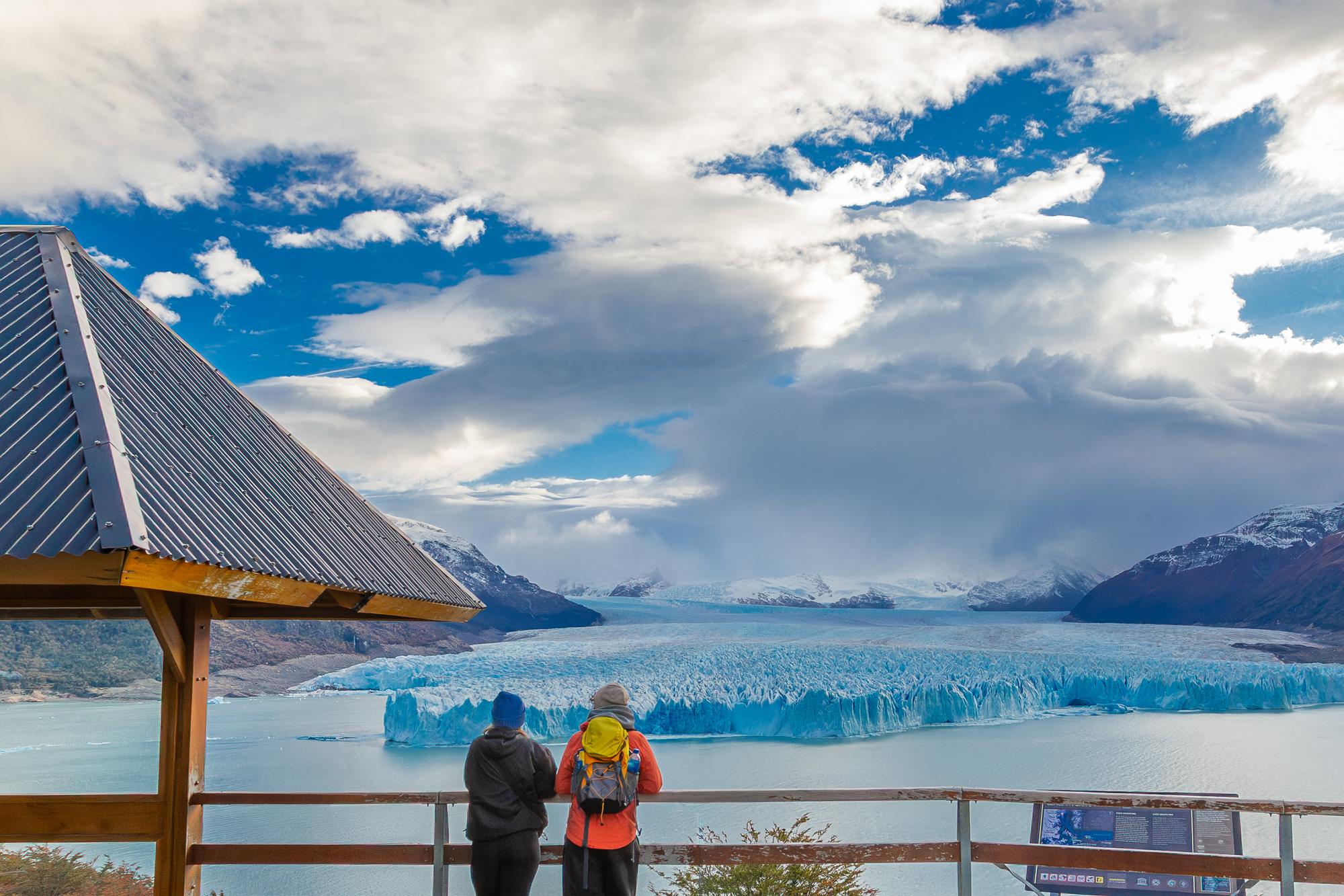 Una pareja contempla el glaciar Perito Moreno desde un mirador, bajo un cielo de nubes