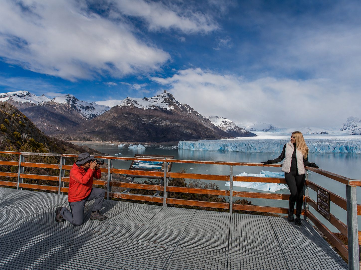 Un fotógrafo arrodillado retrata a una viajera en las pasarelas, con el glaciar detrás